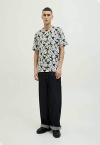 Black and white floral short-sleeve shirt with a loose fit, paired with wide dark denim pants and chunky black shoes, standing against a light background.