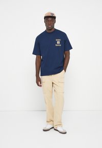Mørkeblå bomulds t-shirt med gul tryk, der siger "Jorden" og "Universitet". Beige cargo bukser med flere lommer. Hvide og grå sneakers.