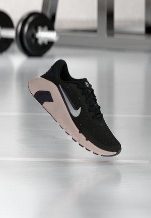 Baskets noires Nike avec logo swoosh blanc flottant au-dessus du sol d’une salle de sport, haltères flous en arrière-plan.