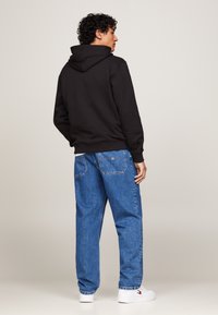 Felpa nera con cappuccio e vestibilità rilassata, abbinata a jeans in denim blu. Il modello è rivolto di spalle, mettendo in evidenza i dettagli posteriori dell'abbigliamento.