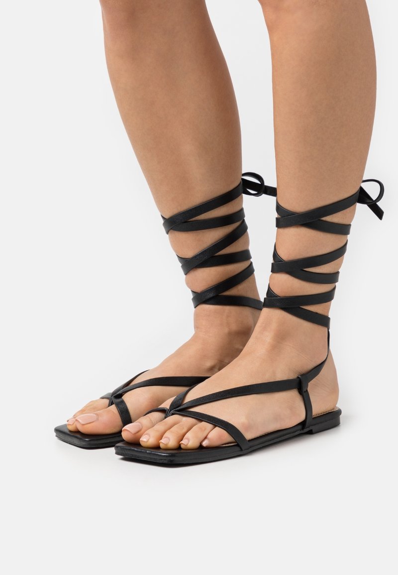 Pieds portant des sandales plates noires avec de fines lanières qui entourent les chevilles et le bas des mollets, nouées en nœuds à l’arrière.