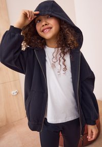 Sweat à capuche noir zippé avec une capuche ample, des poignets côtelés et des poches avant, porté sur un t-shirt blanc et associé à des leggings noirs.