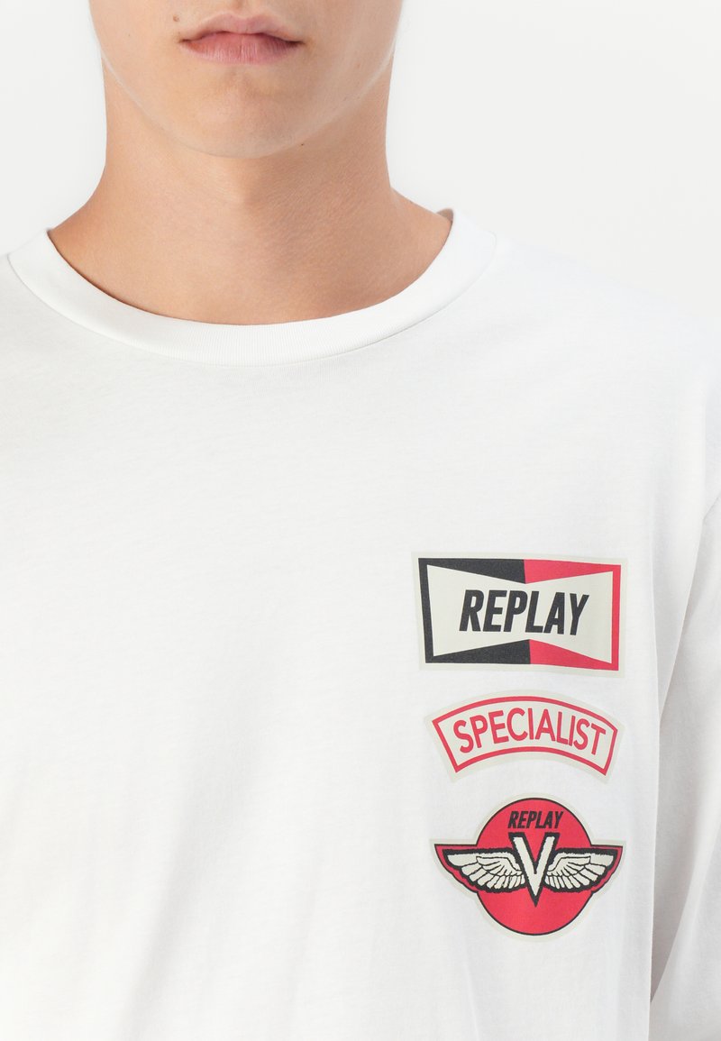 Man in een wit shirt met drie rode, zwarte en witte patches met de teksten "REPLAY," "SPECIALIST," en een gevleugeld "V" logo.