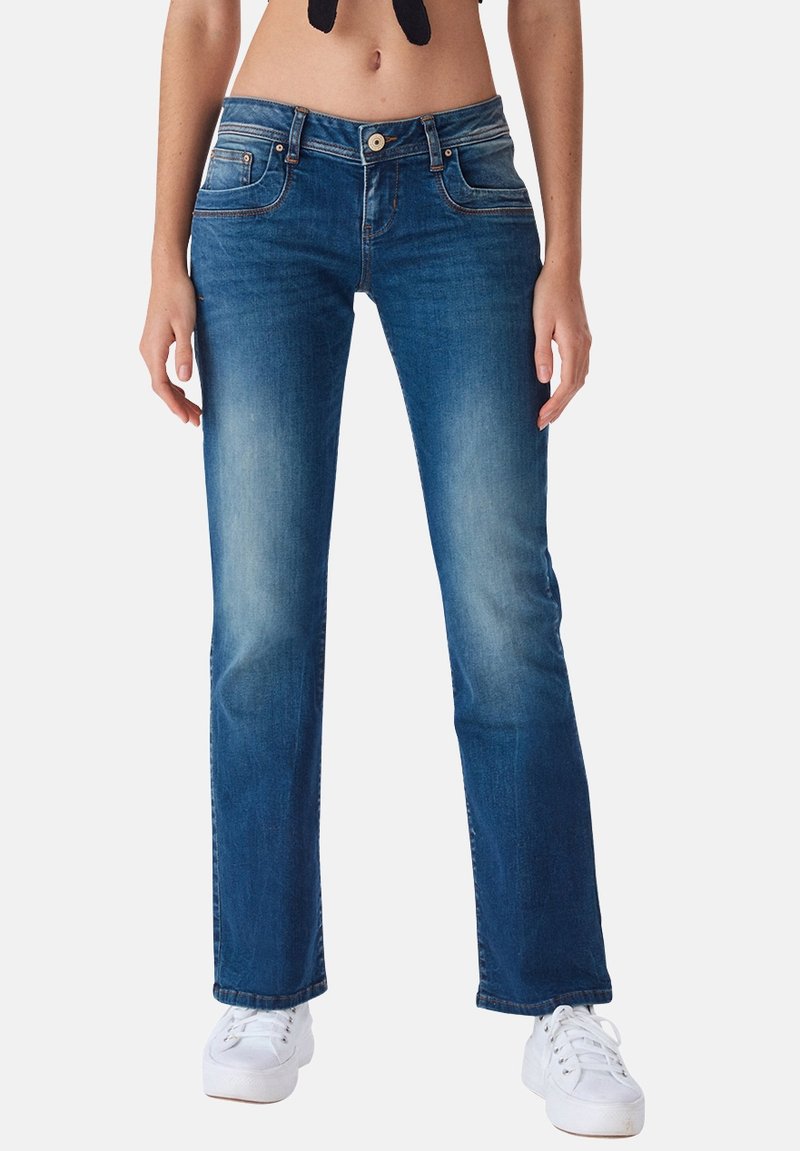 Jeans de denim azul con pierna recta, cierre de botón y bolsillos frontales. Desgaste ligero a lo largo de los muslos y un dobladillo limpio. Zapatillas blancas abajo.