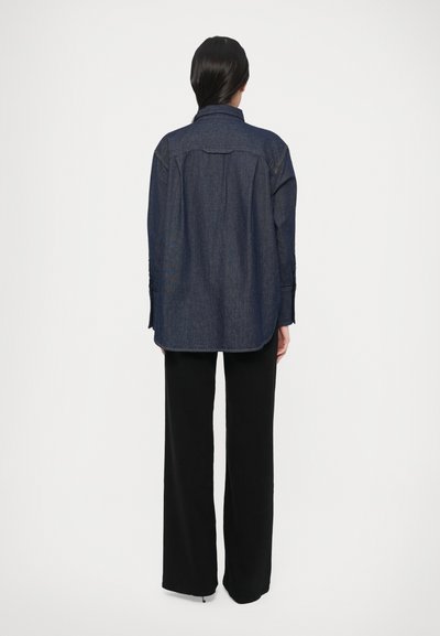Džínová košeľa s voľným strihom, tmavomodrej farby a s detailmi na gombíky, v páre s čiernymi širokými nohavicami. Má štandardný golier a zadný diel s klopou.