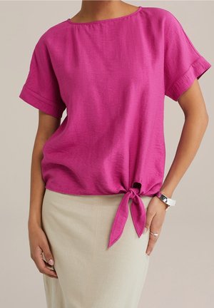 Femme portant un chemisier rose vif à manches courtes noué à la taille, associé à une jupe beige, une bague argentée et un bracelet argenté.