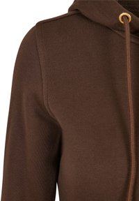 Gros plan sur l'épaule et la poitrine d'un sweat à capuche en tricot marron avec cordon de serrage visible et œillet en métal.