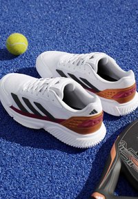 Chaussures de sport blanches avec des rayures noires, une balle de tennis jaune et une raquette sur une surface sportive bleue texturée.