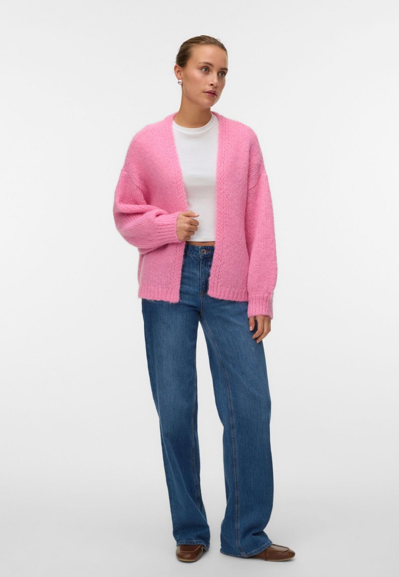 Γυναίκα που στέκεται φορώντας ένα ροζ χοντρό ζακ έτα, λευκή εφαρμοστή μπλούζα, μπλε τζιν παντελόνια με φαρδιά πόδια και καφέ παπούτσια μπροστά σε απλό λευκό φόντο.