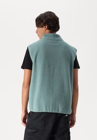 Vesta din polar de culoare teal deschis, cu guler înalt și design fără mâneci, având un finisaj texturat și un tiv posterior puțin mai lung.