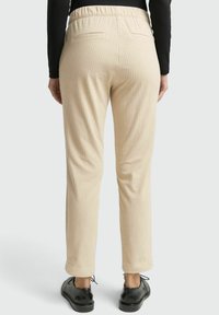 Beige geribbelde broek met elastische tailleband en achterzakken, gedragen met zwarte schoenen en een zwart top met lange mouwen, gezien van achteren.