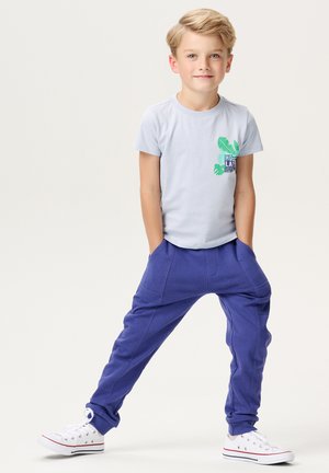 Lichtblauwe katoenen t-shirt met een groene bladerenafbeelding, gecombineerd met paarse joggingsbroek met voorzakken en taps toelopende boorden. Witte sneakers.