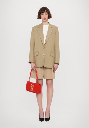 Beige oversized blazer med to forlommer, parret med beige shorts. Modellen holder en rød læderhåndtaske med gyldne bogstaver. Sorte hæle.