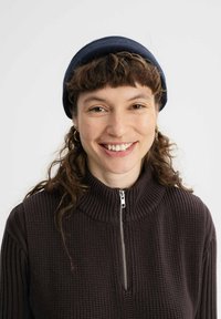 Donkerbruine geribde trui met een hoge kraag en een zilveren rits, gecombineerd met een marineblauwe beanie. Krullend haar en ronde oorbellen zijn zichtbaar.