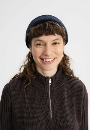 Pull à côtes marron foncé avec un col haut et une fermeture éclair argentée, associé à un bonnet bleu marine. Des cheveux bouclés et des boucles d'oreilles rondes sont visibles.