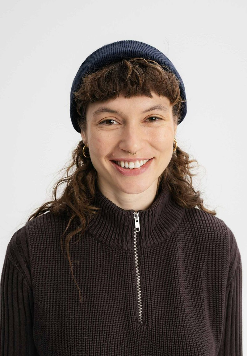 Donkerbruine geribde trui met een hoge kraag en een zilveren rits, gecombineerd met een marineblauwe beanie. Krullend haar en ronde oorbellen zijn zichtbaar.