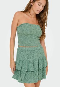 Mujer vestida con un top corto texturizado sin tirantes verde y una falda floral en capas a juego, de pie contra un fondo claro y liso.
