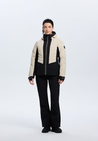 Beige und schwarze Steppjacke mit Kapuze, mit Zickzack-Nähten, Reißverschluss vorne und elastischen Bündchen. Kombiniert mit schwarzen Hosen.
