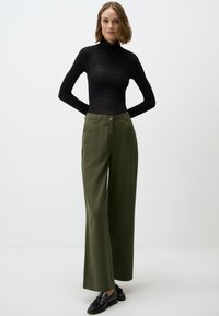 Maglione nero a maniche lunghe con collo alto abbinato a pantaloni olive verdi a vita alta e a gamba larga, dotati di due tasche frontali e una texture liscia. Scarpe nere con nappine.