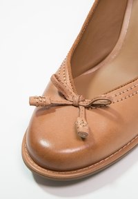 Chaussure en cuir brun clair avec un bout arrondi, décorée d'un nœud et de surpiqûres le long du bord. Texture lisse dans l'ensemble.