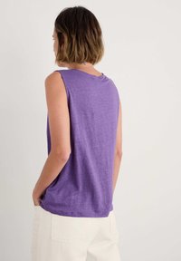 Purple sleeveless top with a casual fit and round neckline, featuring soft, lightweight texture and a curved hemline. Paired with white trousers.