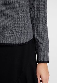 Pull gris tricoté avec une texture côtelée, un ourlet contrastant bleu marine et des manches longues. Le tissu est épais et chaud avec un design structuré.