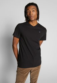 Black v-neck t-shirt made from soft fabric, featuring short sleeves and a subtle logo detail on the chest. Paired with light brown corduroy pants.