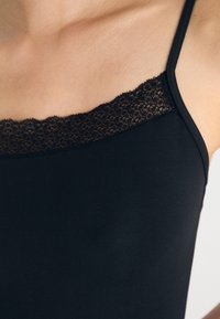 Svart tanktop med tynne stropper, med en blondeaksecent ved utringningen. Stoffet er glat med en tettsittende silhuett.