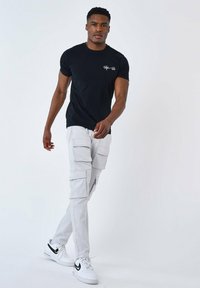 Camiseta negra de manga corta con logo, combinada con pantalones cargo grises claros que tienen múltiples bolsillos, y zapatillas blancas con acentos en negro.