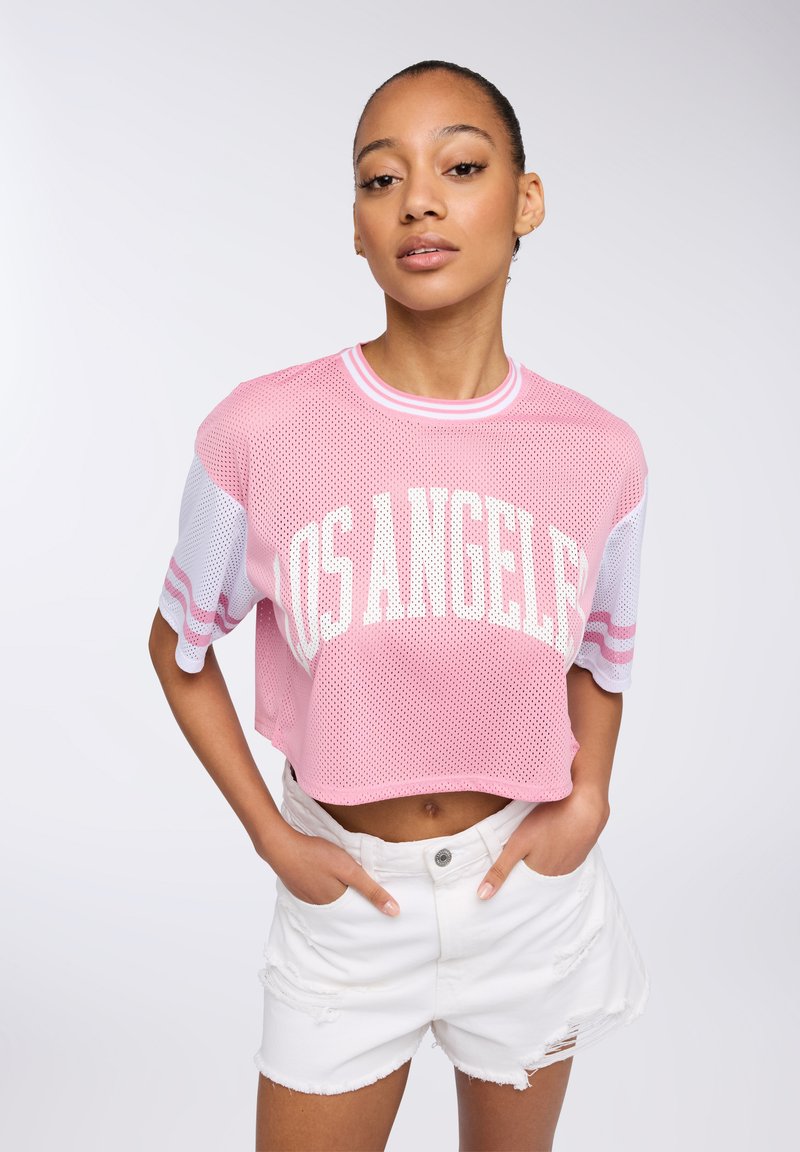 Maglietta cropped in rete rosa con la scritta "LOS ANGELES" in bianco, con maniche a righe bianche e rosa. Abbinata a pantaloni corti bianchi strappati.