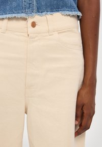 Pantalones de mezclilla color crema con cintura alta, que cuentan con un bolsillo frontal y un cierre de botón metálico. Tejido texturizado con costuras visibles.