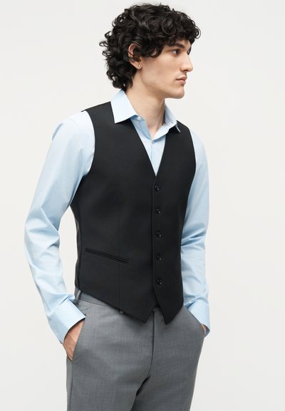 Άντρας με ανοιχτό μπλε πουκάμισο, μαύρο γιλέκο με κουμπιά και γκρι παντελόνια, χέρια στις τσέπες, κοιτάζει προς τα δεξιά σε απλό φόντο.
