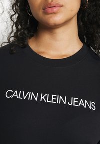 Felpa nera con scollatura a costine, presenta la scritta bianca "CALVIN KLEIN JEANS." Tessuto morbido, design casual, aderente attorno al collo.