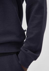 Felpa navy con un tessuto texturizzato, dotata di polsini arrotolati e un piccolo logo ricamato sul petto. Design minimalista con branding discreto.