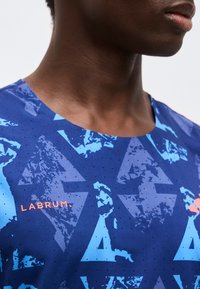 Κοντινό πλάνο ενός ατόμου που φορά μπλε πουκάμισο με αφηρημένα τριγωνικά σχέδια και τη λέξη "LABRUM" τυπωμένη πορτοκαλί στο στήθος.