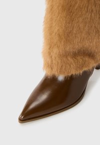 Stivale alla caviglia in pelle marrone con una soffice finitura in pelliccia texturizzata color tan sulla parte superiore, caratterizzato da una superficie lucida e un tacco piccolo.