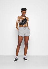 Maglietta a maniche lunghe marrone tie-dye, pantaloni corto grigi con coulisse e marsupio nero; abbinati a sneakers bianche e nere.