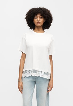 Γυναίκα με σγουρά μαλλιά φοράει μια φαρδιά λευκή μπλούζα με δαντελένια διακόσμηση πάνω από ανοιχτό μπλε τζιν, στέκεται μπροστά σε απλό λευκό φόντο.