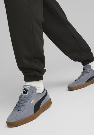 Person trägt graue Puma Wildledersneaker mit schwarzen Streifen, weißen Socken und schwarzen Jogginghosen vor einem einfarbigen Hintergrund.