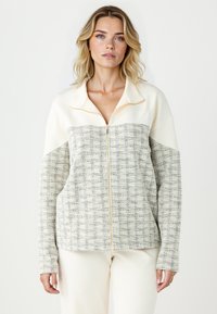 Veste zippée avec une partie supérieure crème et une partie inférieure beige texturée, arborant un motif à carreaux, des manches longues et des détails de fermeture éclair dorés.