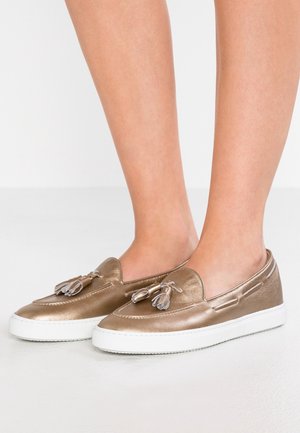 Metalliske bronze slip-on loafers med kvaster, båret på bare fødder, med hvide gummisåler, vist mod en enkel lys baggrund.