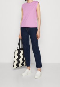 Růžový bezramínkový top, tmavě modré slim-fit džíny a bílé slip-on obuv. Drží černobílou prošívanou tote tašku s vlnitým vzorem.