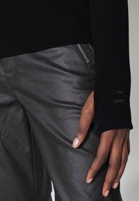 Black ribbed long-sleeve top paired with sleek black leather pants featuring a side zipper. The top has a subtle brand logo on the sleeve.
