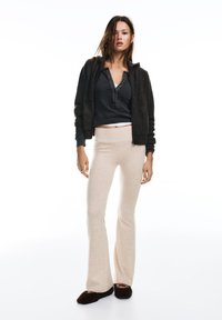 Szürke cipzáras kapucnis pulóver, fekete henley felső, bézs bővülő szárú nadrág és sötét papucsok. Puha anyagok, laza szabás és semleges színpaletta.