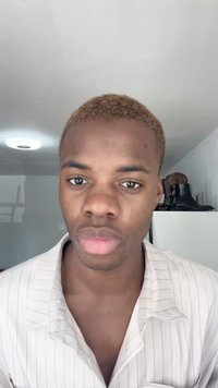 Sujeito do sexo masculino com cabelo curto e loiro claro, vestindo uma camisa branca com riscas. O fundo apresenta uma parede branca e calçado de cor escura.