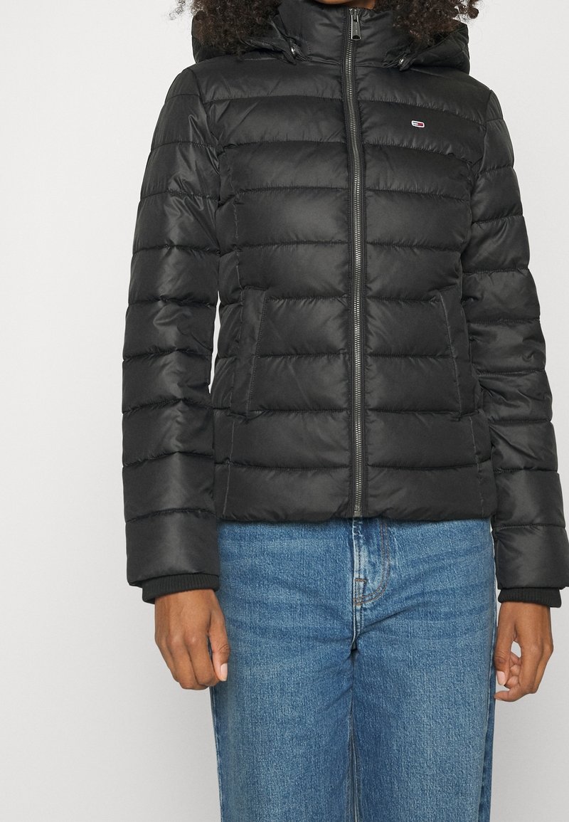 Personne portant une doudoune matelassée noire avec fermeture éclair frontale et capuche, associée à un jean en denim bleu, debout devant un fond uni.