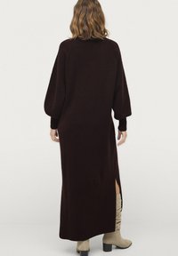 Lange bordeauxrode gebreide jurk met ballonsleeves, zijsplitten en ribgebreide manchetten en onderrand. De textuur van de stof voegt warmte en diepte toe.