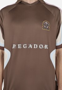 Rjava športna majica s ovratnikom, ki vsebuje logo "PEGADOR" in teksturirano blago. Omake v svetlo modri barvi na rokavih.