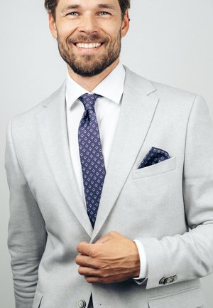 Naeratav mees kohandab helehallit ülikonnajopet, kandes valget särk ja sinist mustrilist lipsu sobiva taskurätikuga.