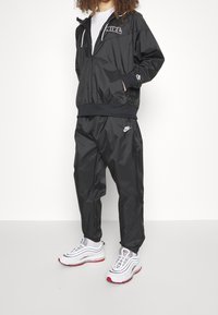 Svart nylonträningsoverall med en jacka med dragkedja och logotyp samt matchande byxor. Bärs med vita och röda träningsskor.